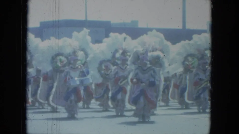 1966 Milwaukee home-movie frame showing a line of elaborately dressed parade performers with white feathered costumes.
