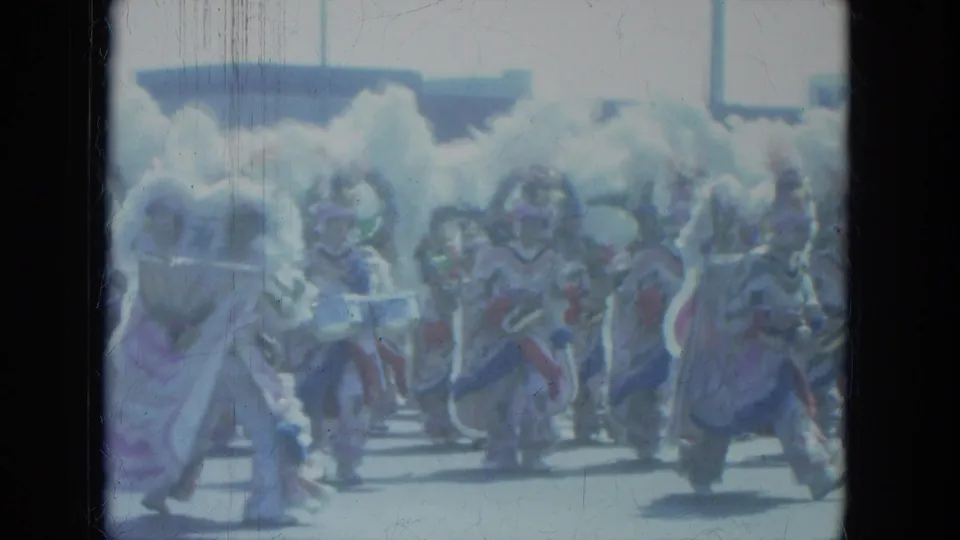 1966 Milwaukee parade frame showing costumed performers in white and purple feathered outfits crossing the camera's view.