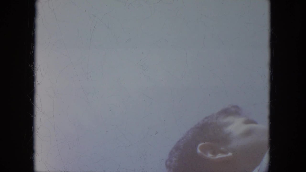 Brief 1966 Milwaukee home-movie frame showing a spectator's head at the lower right against a pale, overexposed background.