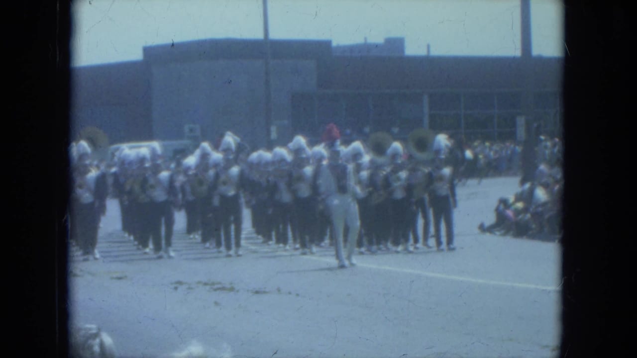 1966 Milwaukee parade frame showing a marching band in formation with a conductor in front and spectators along the curb.