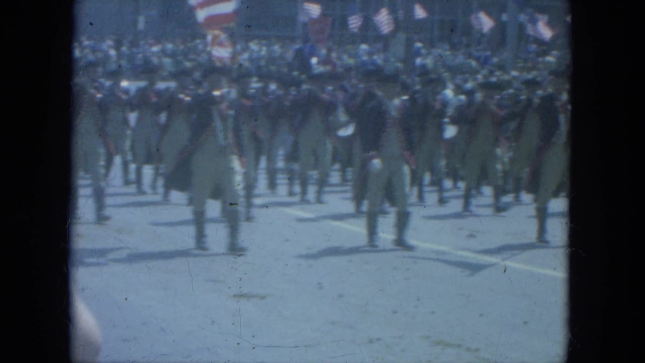 Men in colonial-style parade uniforms continue marching in formation on a Milwaukee street in 1966 home-movie footage.