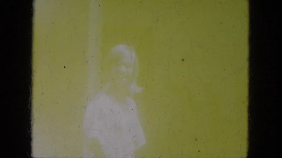 In 1966 Milwaukee home-movie footage, a young blond girl in a light floral shirt or dress smiles toward the camera indoors.
