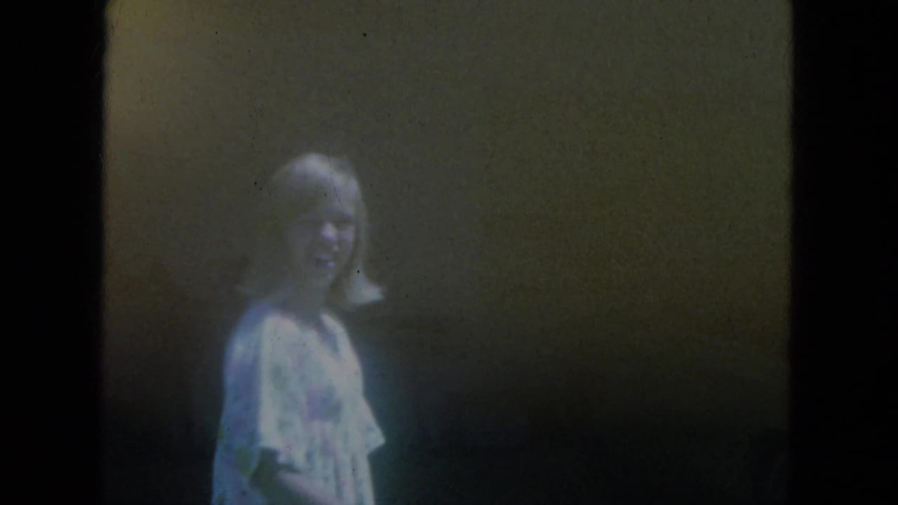 In 1966 Milwaukee home-movie footage, a woman in a light dress smiles toward the camera in a dark interior doorway.