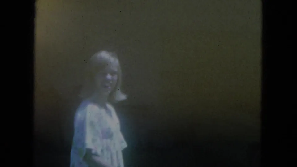 In 1966 Milwaukee home-movie footage, a woman in a light dress smiles toward the camera in a dark interior doorway.