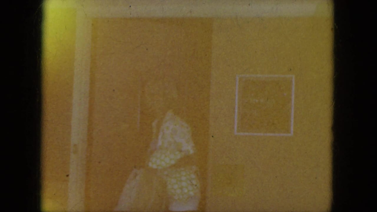 The woman in a light polka-dot dress moves through a warm-toned interior doorway, with a framed picture on the wall.