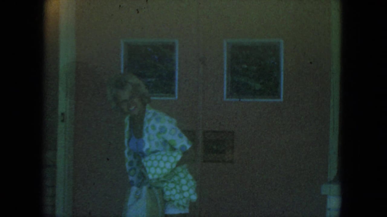 Adult woman in 1966 Milwaukee home-movie footage stands near a wooden door and wall, partly obscured by shadow and film grain.