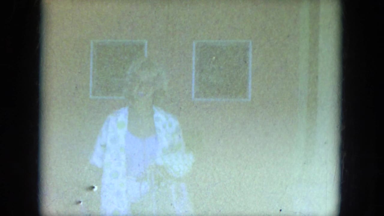 Overexposed 8mm frame shows the woman in a light patterned garment near a wall and doorway in Milwaukee home-movie footage.