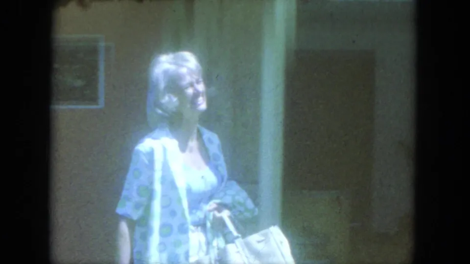 Blonde adult woman in a patterned housecoat walks past a doorway while carrying a light-colored bundle in 1966 Milwaukee footage.