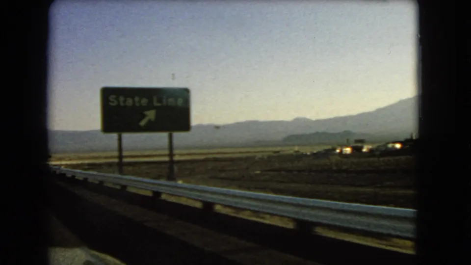 1966 Nevada road view with a green State Line sign, metal guardrail, desert flats, and distant mountains.