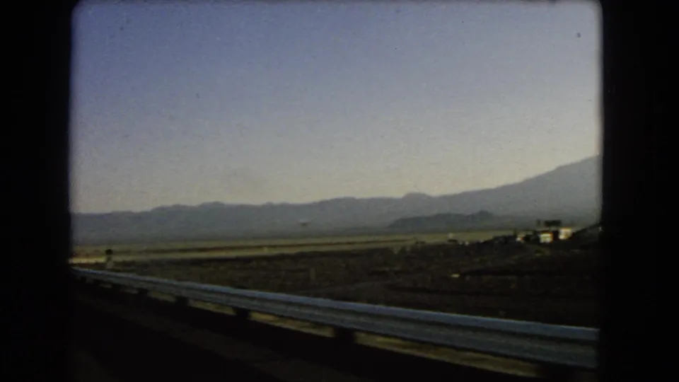 1966 Nevada roadside view showing desert flats, a metal guardrail, distant mountains, and small structures near the horizon.