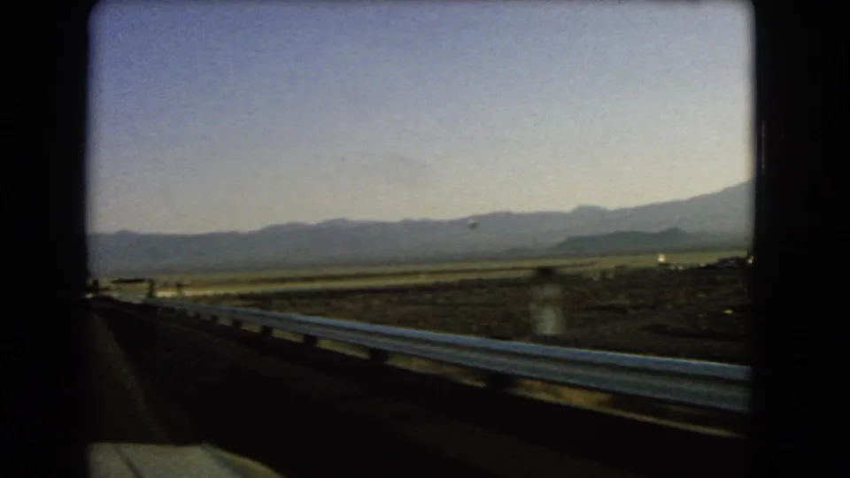 Moving-car view along a Nevada desert highway, with guardrail leading toward open flats and a mountain range.