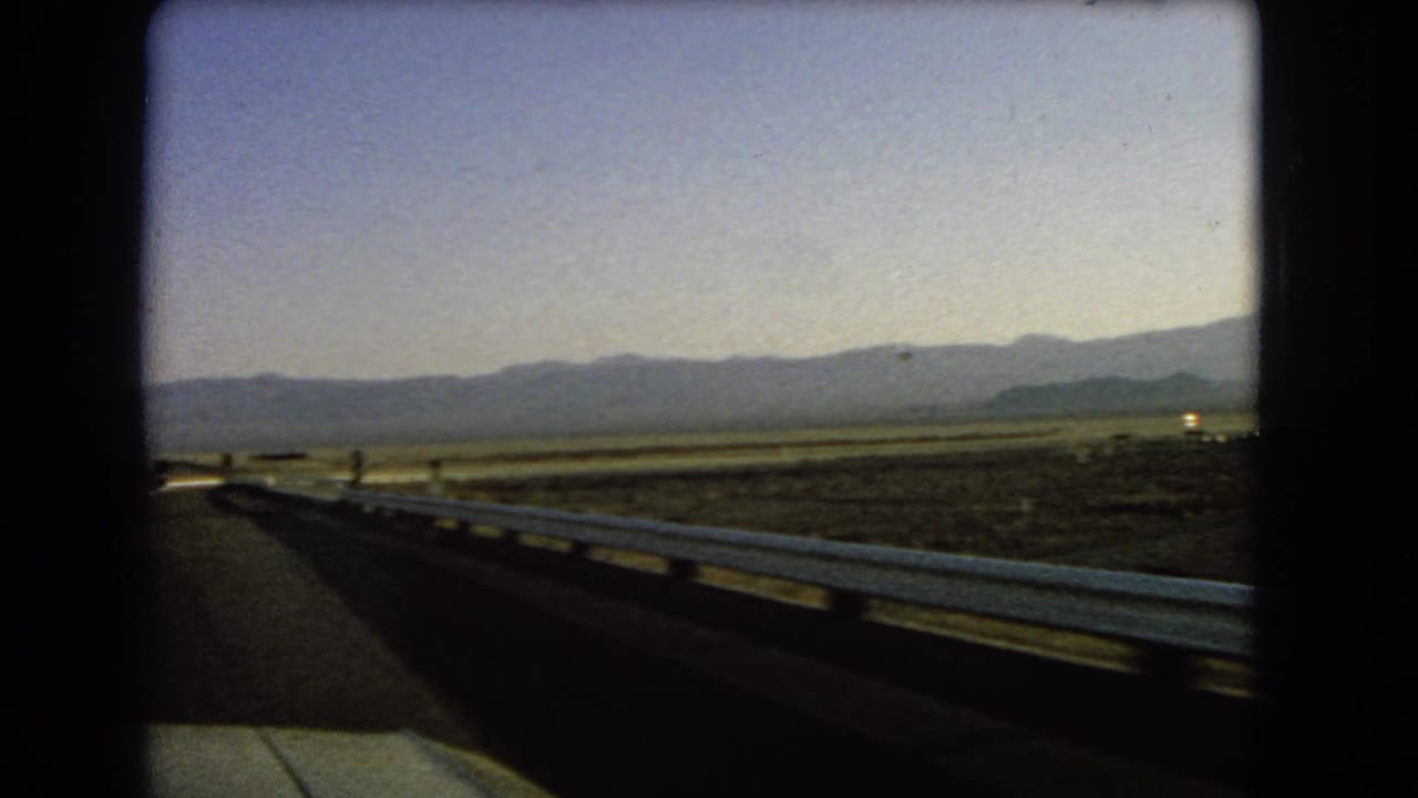 1966 Nevada 8mm view from a moving vehicle along a highway with a white guardrail, open desert land, and far mountains.