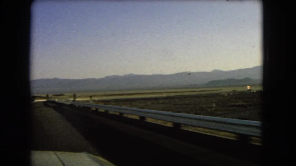 1966 Nevada 8mm view from a moving vehicle along a highway with a white guardrail, open desert land, and far mountains.