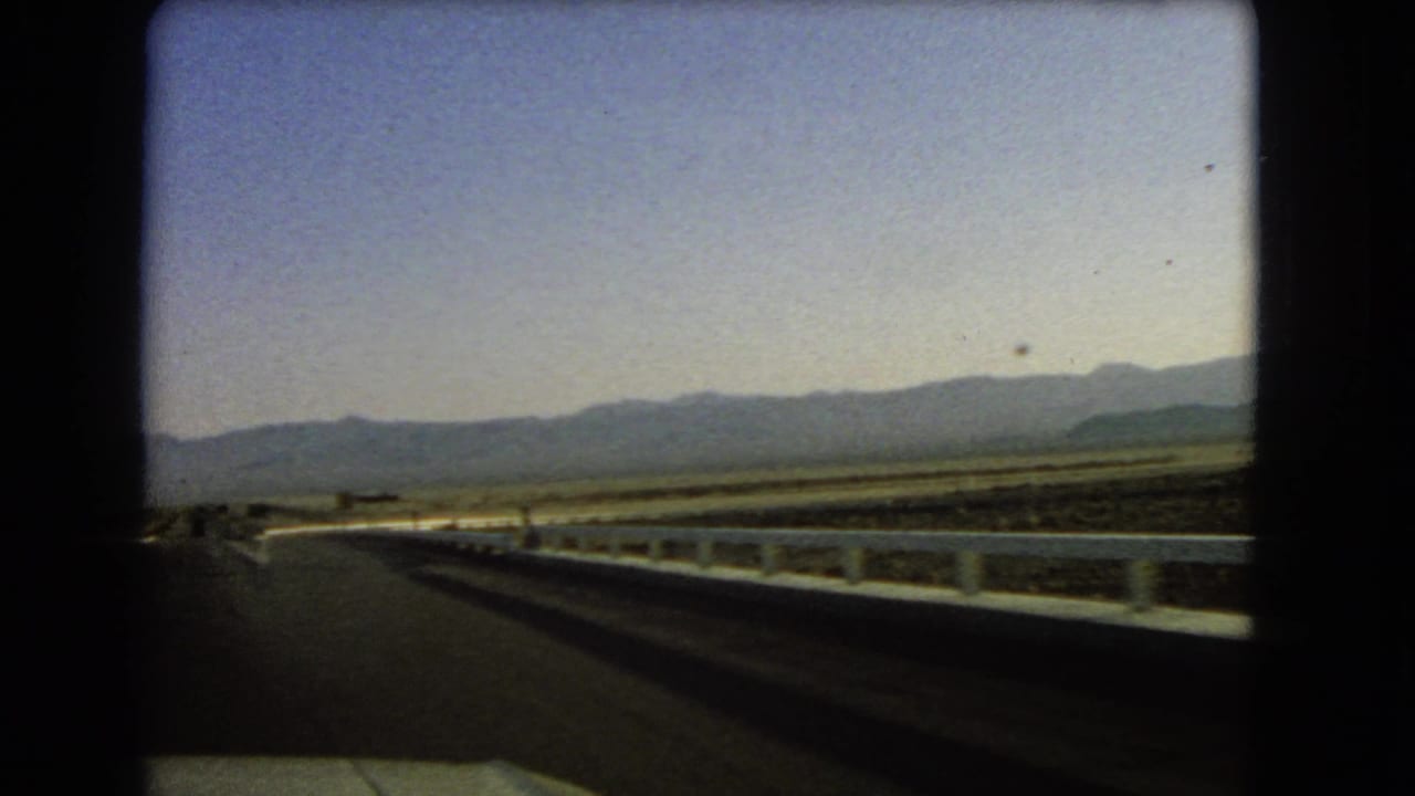 Moving-vehicle view on a Nevada highway in 1966, showing a guardrail, flat desert terrain, and a hazy mountain range.
