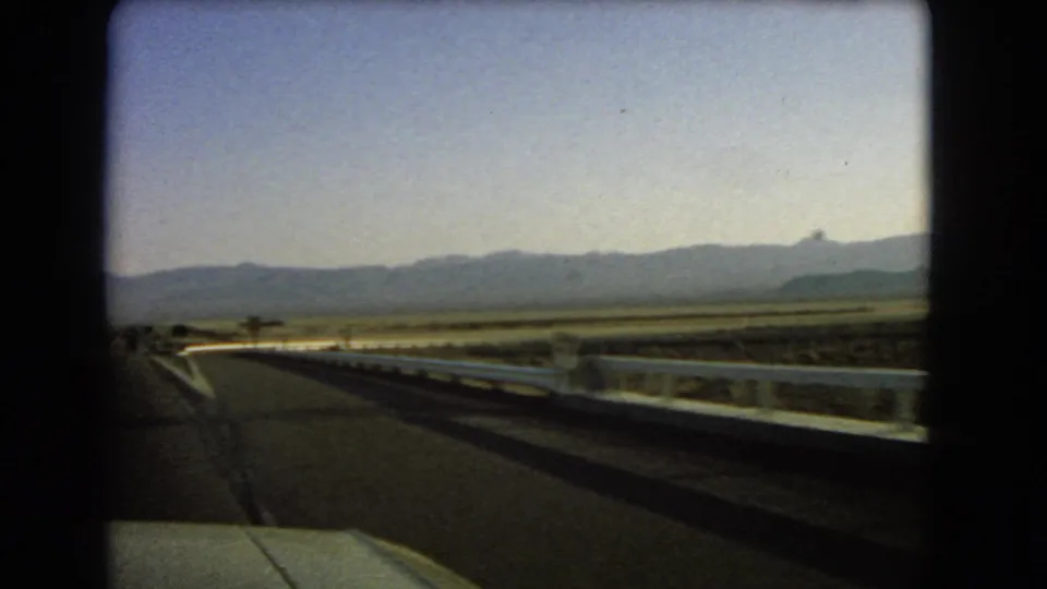 1966 home-movie frame from Nevada showing a roadside guardrail, paved highway, desert scrub, and distant mountain ridge.