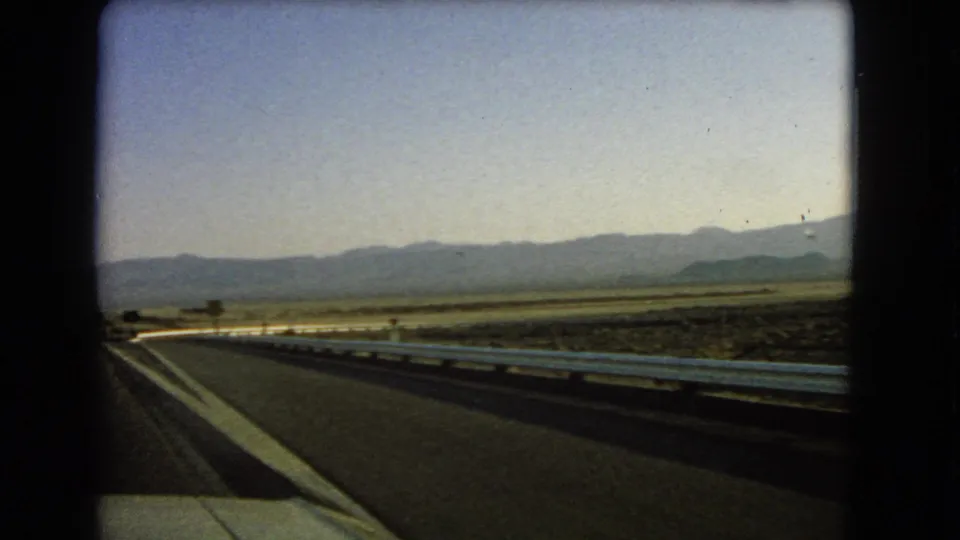 Nevada desert highway view from 1966 8mm footage, with the road edge, white guardrail, broad flats, and far mountains.