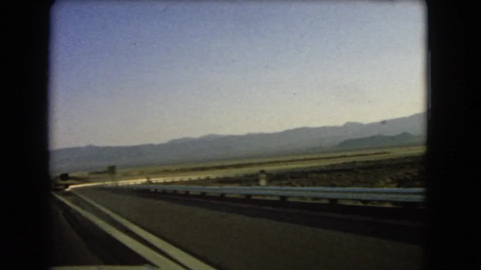 1966 Nevada 8mm passenger-view footage of an empty desert highway, roadside guardrail, open plain, and distant mountains under a pale sky.