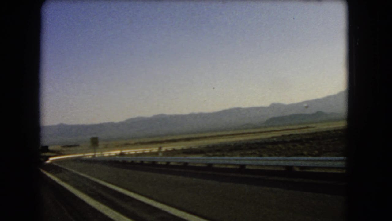Roadside sign and guardrail pass along an empty highway with low mountains beyond.
