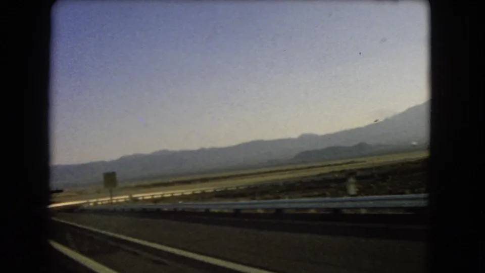 1966 Nevada home-movie frame from a moving car, showing the road edge, guardrail, roadside sign, desert flats, and mountains.