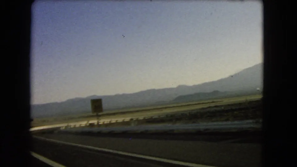 End frame of the 1966 Nevada road sequence, with a roadside sign near the guardrail and mountains across the desert plain.