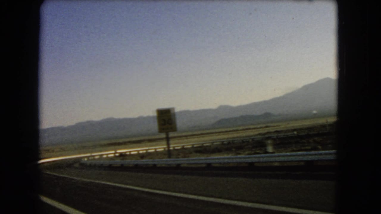 Desert highway curve with guardrail, distant mountains, and a roadside speed sign.