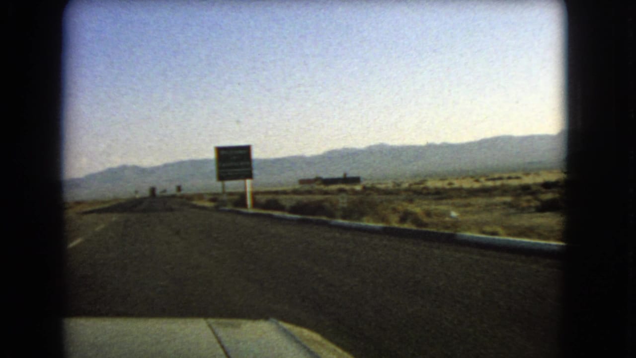 Straight highway approaching a roadside sign with desert scrub and mountains beyond.