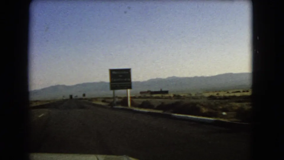 Nevada-to-California road-trip view from 1966 showing a car approaching a green state welcome sign beside a desert highway.