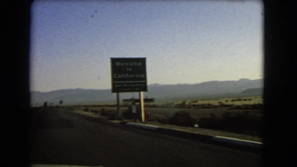 1966 8mm frame showing a Welcome to California roadside sign near the Nevada border, with desert scrub and mountains behind it.