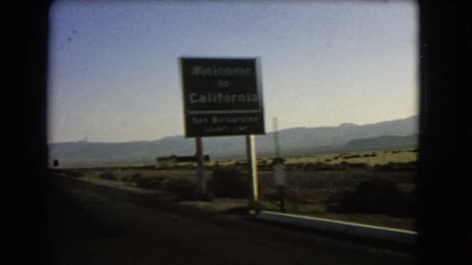1966 8mm road-view frame showing a Welcome to California sign at the San Bernardino County line with desert mountains beyond.