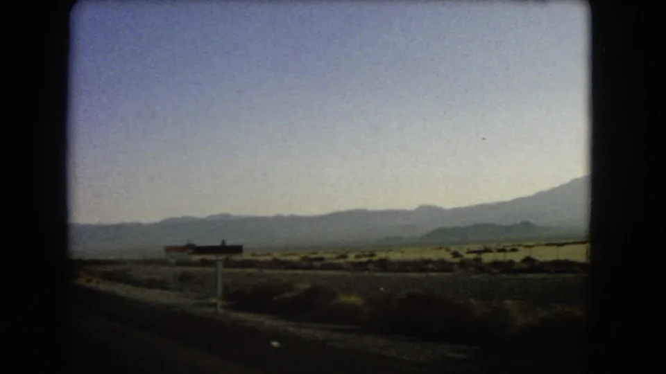 1966 8mm car-window view of open desert terrain near the California border, with low mountains and roadside signs in the distance.