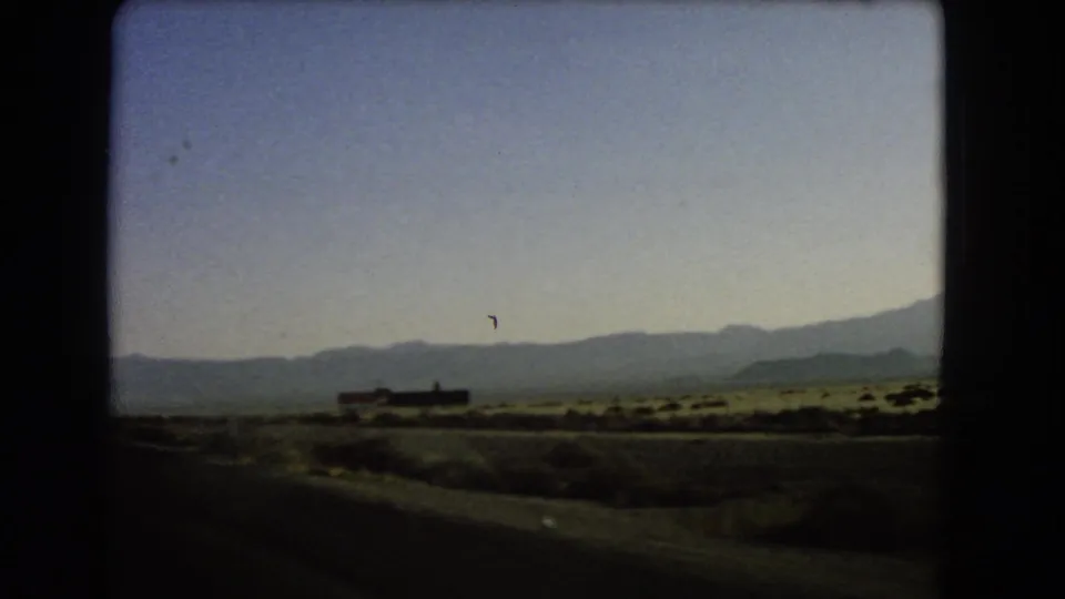 1966 8mm frame from a moving car showing a sparse desert shoulder, distant sign, and mountain range under a clear sky.