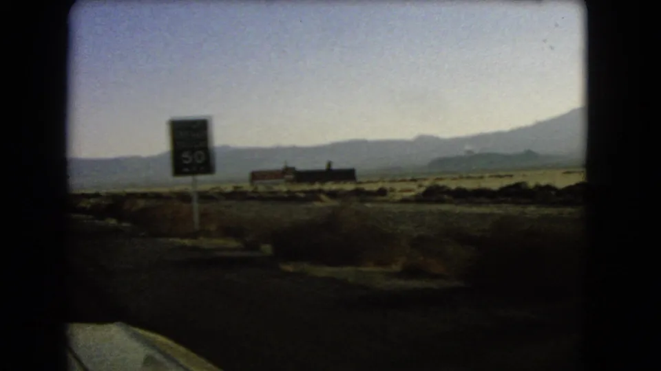 1966 8mm road-view frame showing a truck speed-limit sign by the desert highway, with open scrubland and mountains in the distance.