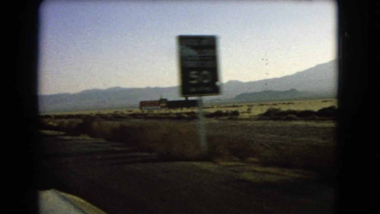 Desert roadside seen from a moving car with a blurred sign and mountains in the distance.