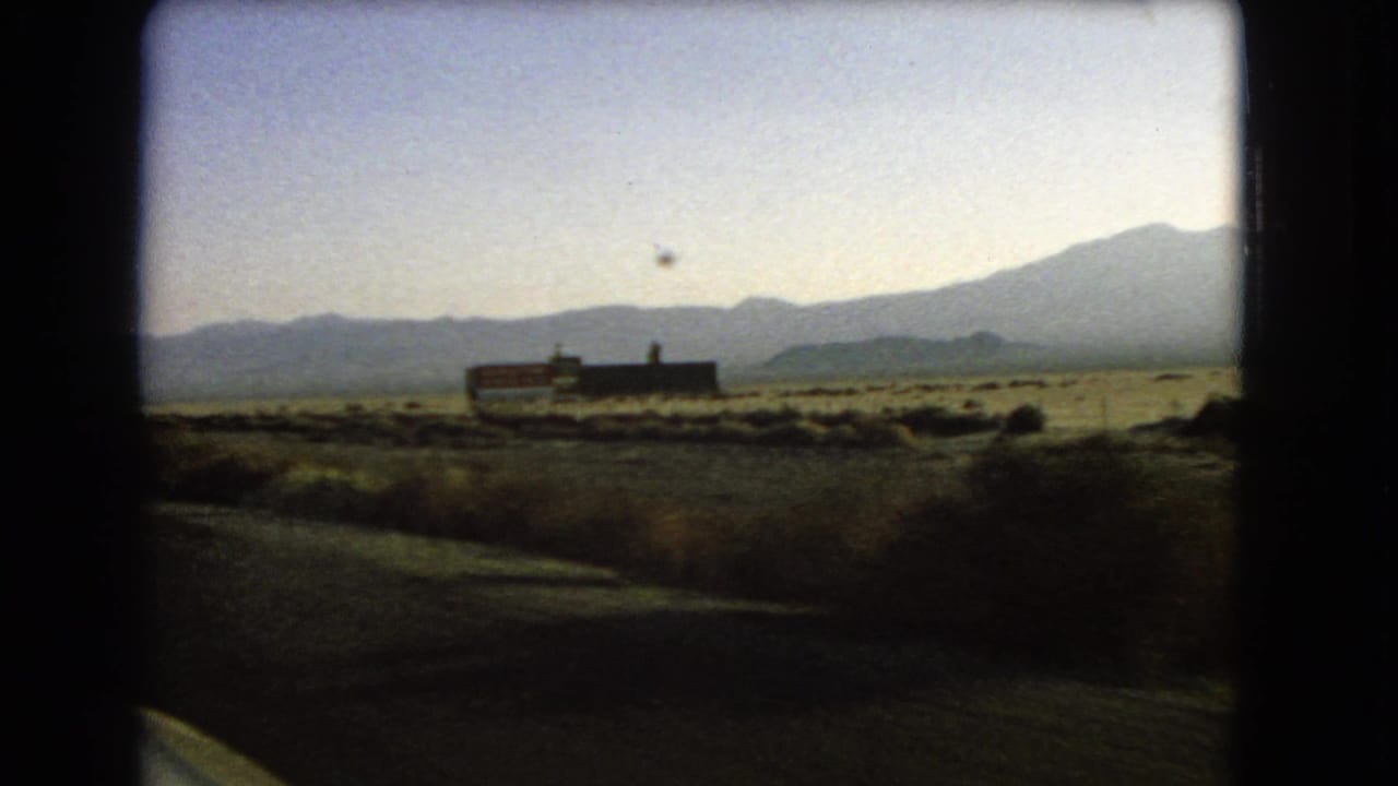 1966 Nevada desert highway view with low roadside signage, dry scrub, and a mountain range beyond the road shoulder.