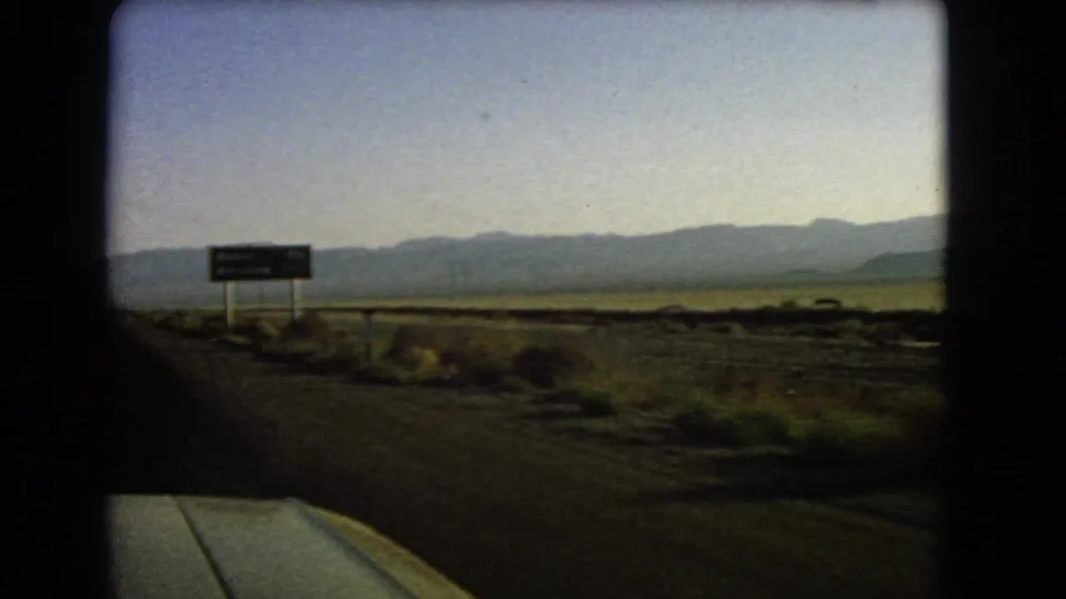 1966 Nevada road footage showing a roadside traffic sign beside desert scrub, with mountains stretching across the horizon.