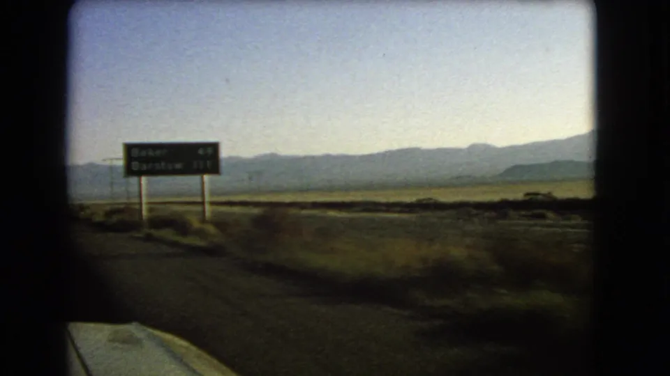 1966 Nevada desert road view showing a highway destination sign, likely for Baker and Barstow per metadata, with mountains beyond.