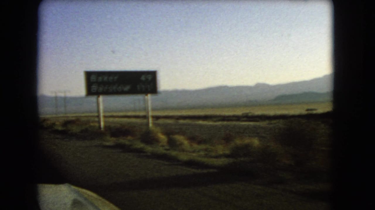 Green highway distance sign beside a desert road with mountains in the background.