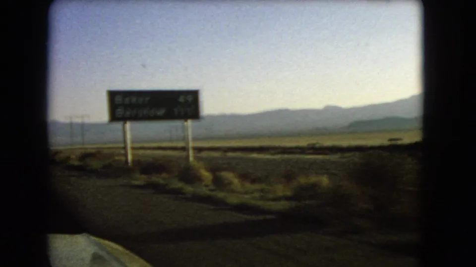 1966 Nevada 8mm frame showing a green highway sign for Baker and Barstow beside open desert terrain and distant mountains, filmed from a moving car.