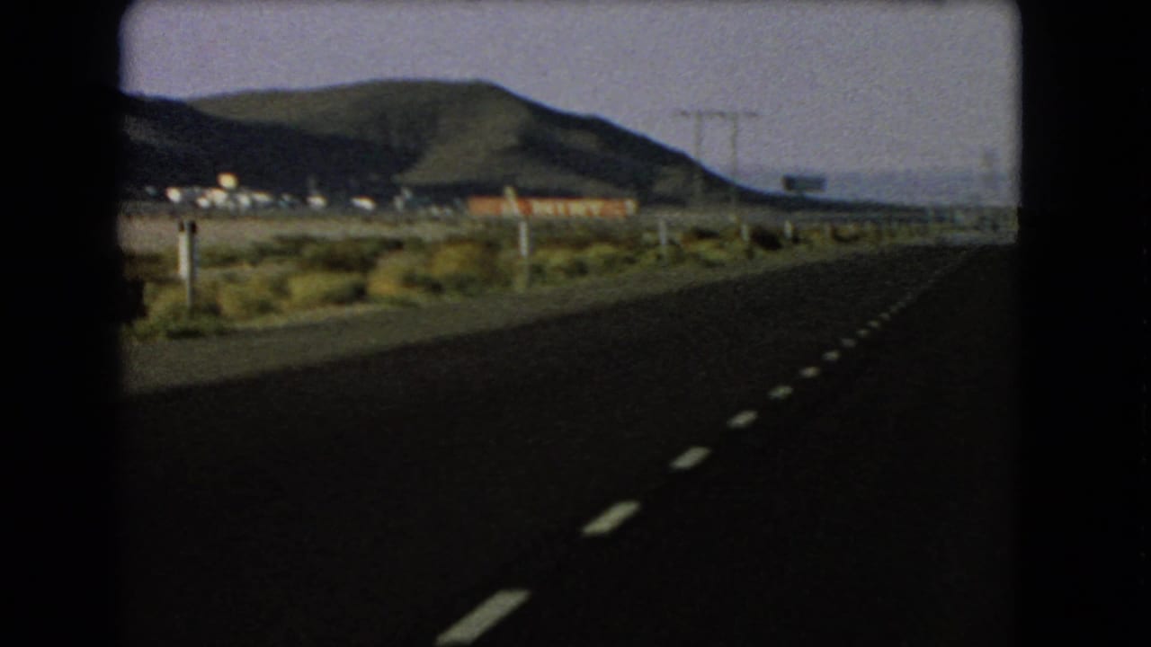 1966 Nevada road-trip frame showing a paved highway, dashed center markings, roadside posts, desert vegetation, utility poles, low buildings, and hills beyond.