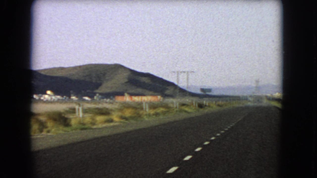 Moving-car 8mm view of a Nevada desert highway with dashed lane markings, roadside buildings, utility poles, scrub, and dark low mountains in the distance.