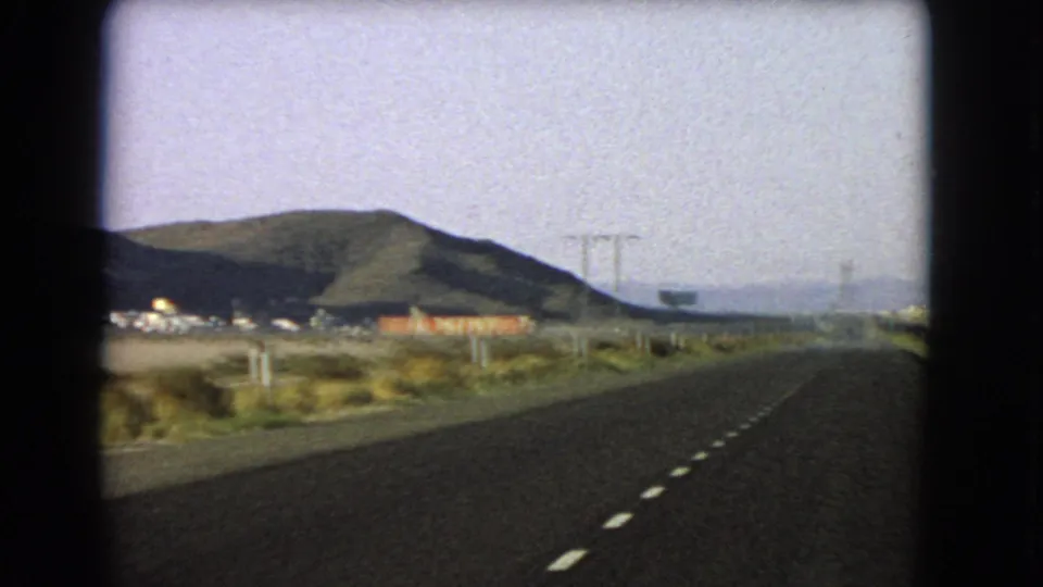 Moving-car 8mm view of a Nevada desert highway with dashed lane markings, roadside buildings, utility poles, scrub, and dark low mountains in the distance.