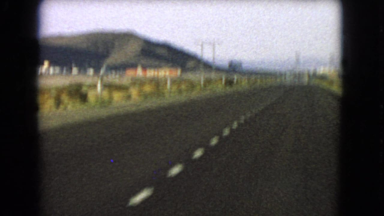 1966 Nevada home-movie view down a paved desert highway with dashed lines, low mountains and scattered roadside structures.