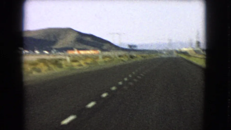 A 1966 Nevada desert highway stretches ahead from a moving car, bordered by scrubland, utility poles and distant buildings.