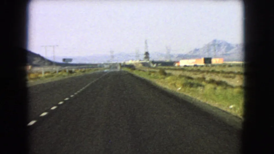 Late-frame 1966 Nevada road view with open highway, roadside sign, utility poles, pale building and distant mountain forms.