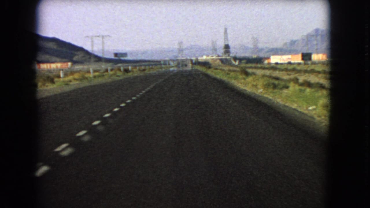 1966 Nevada road-trip view from inside a vehicle, looking down a straight highway toward distant hills and roadside structures.