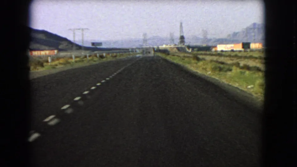 1966 Nevada road-trip view from inside a vehicle, looking down a straight highway toward distant hills and roadside structures.