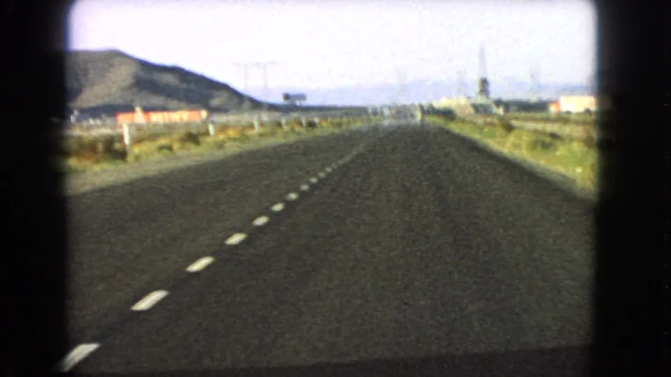 Passenger-view 8mm frame of a Nevada highway in 1966, with dashed road lines, arid shoulders, and distant hills.
