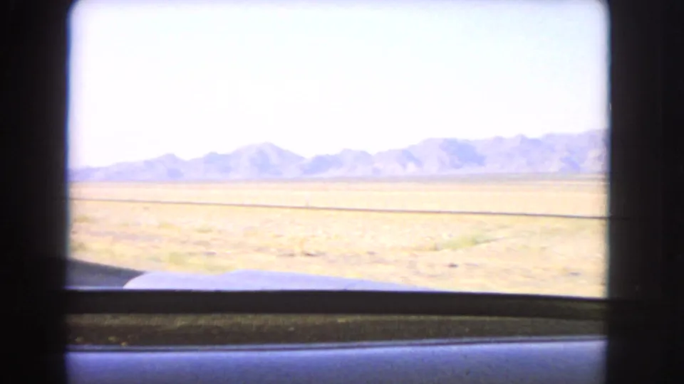 1966 Nevada desert vista from a moving car, with flat open ground, distant mountains, and windshield edge visible.