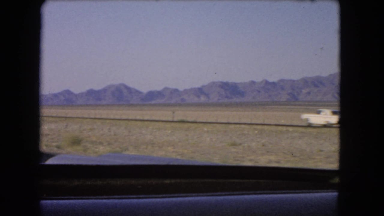 White car on a highway with desert plain and mountains beyond.