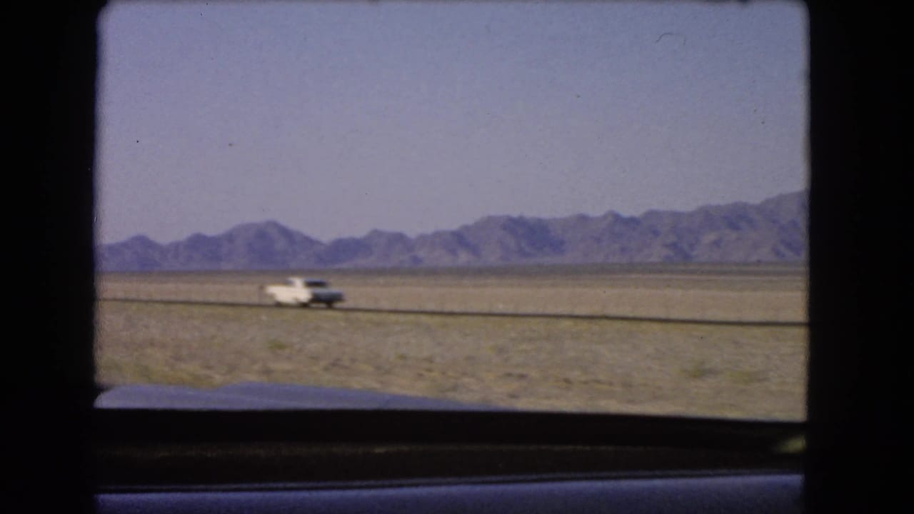 Mid-1960s Nevada highway view with a white car crossing the frame against desert and mountain scenery.